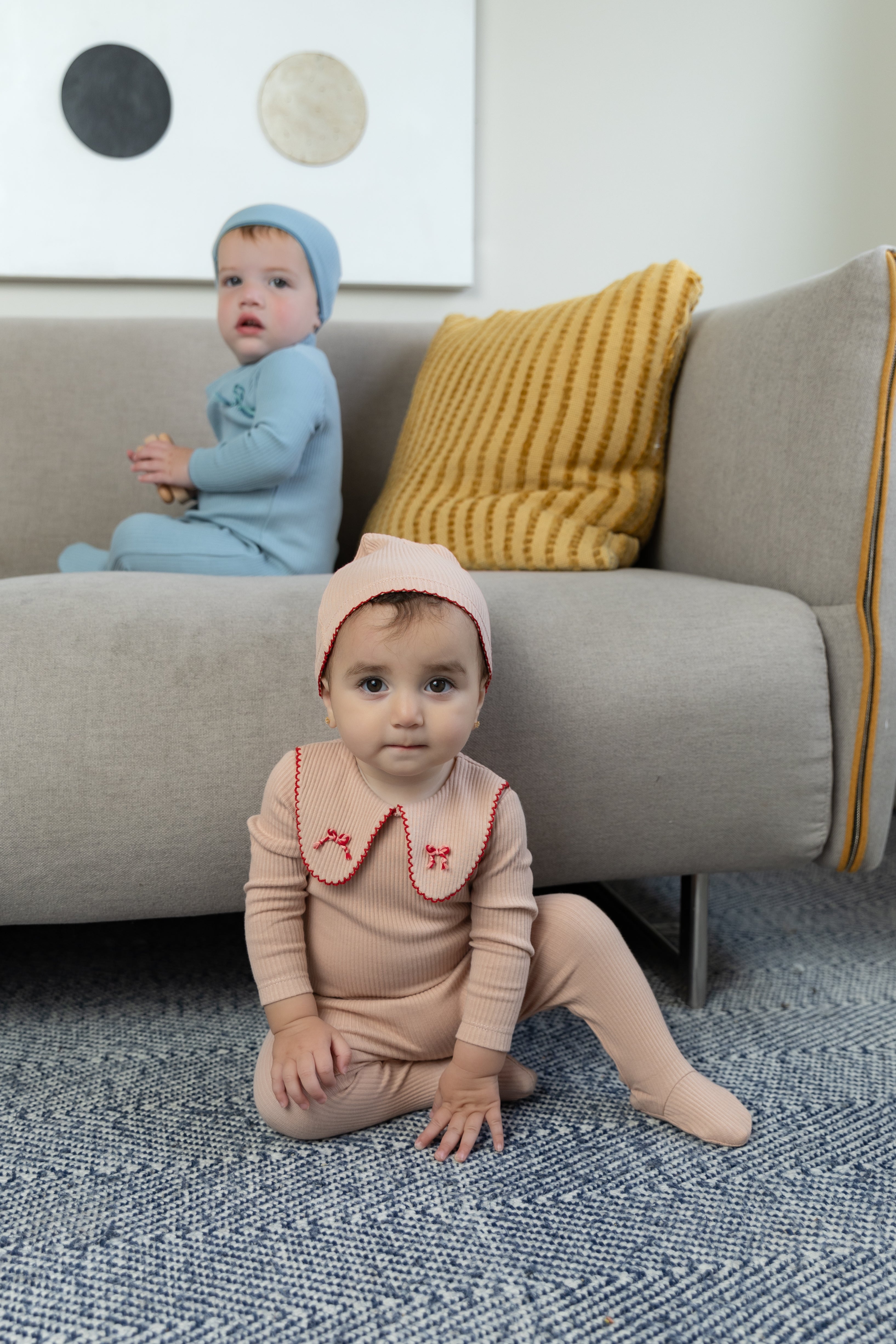 Two babies in colorful outfits sitting on a couch with decorative pillows.